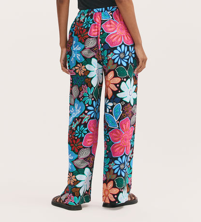 A person wears Eden Blue Floral Crepe Wide Leg Trousers, featuring a vibrant multicolored floral print in blue, pink, red, and white with an elasticated waistband, standing against a plain light background.