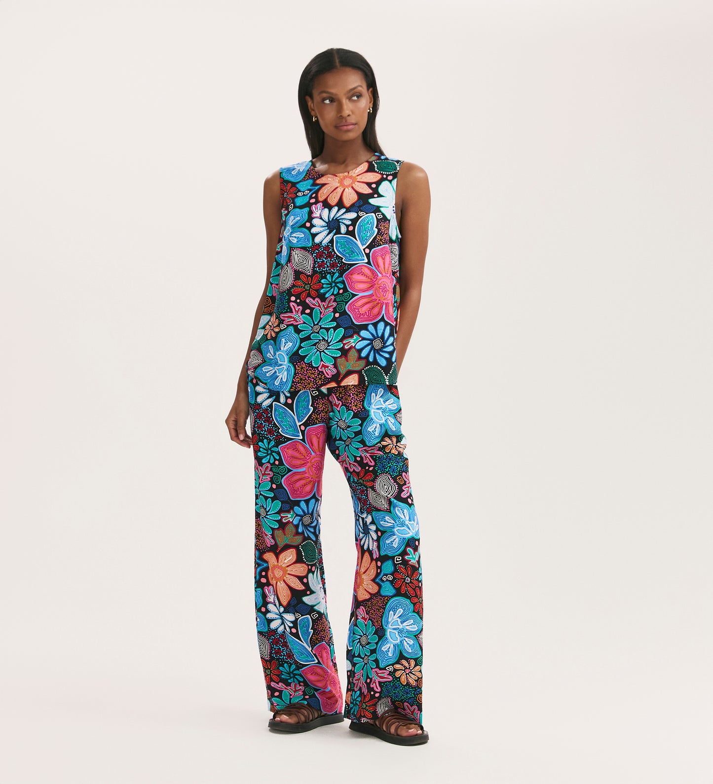 A woman poses against a white backdrop wearing the Eden Blue Floral Crepe Wide Leg Trousers, featuring a bold blue, red, pink, and white floral print on black with an elasticated waistband.