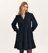 A woman with long blonde hair wears the Verona Navy Corduroy Short Dress, featuring long sleeves, a tiered skirt, and buttoned neckline, standing against a plain light background.