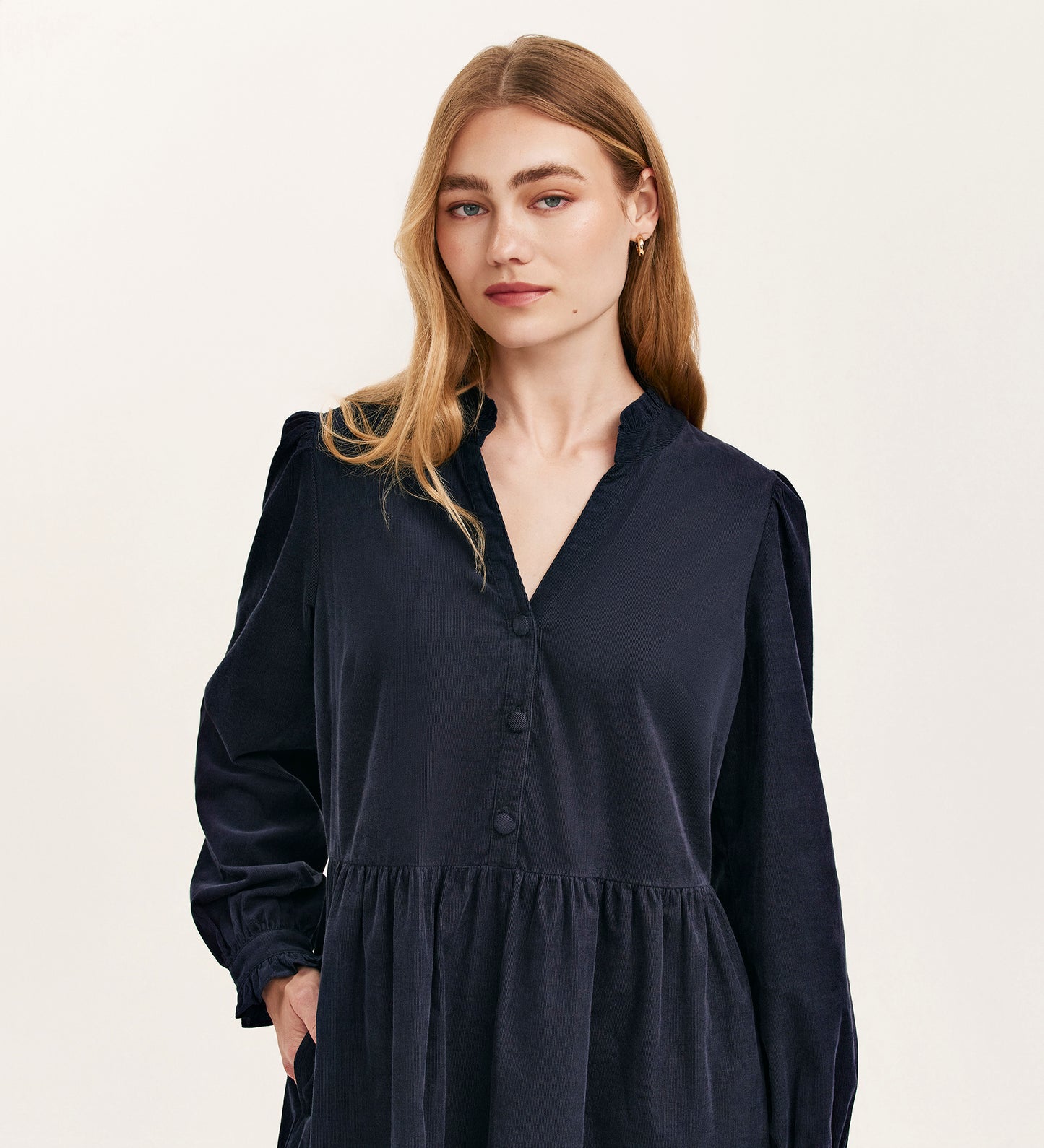 A woman with long blonde hair wears the Verona Navy Corduroy Short Dress, featuring a v-neck and buttons, standing against a plain light background and looking directly at the camera with a neutral expression.