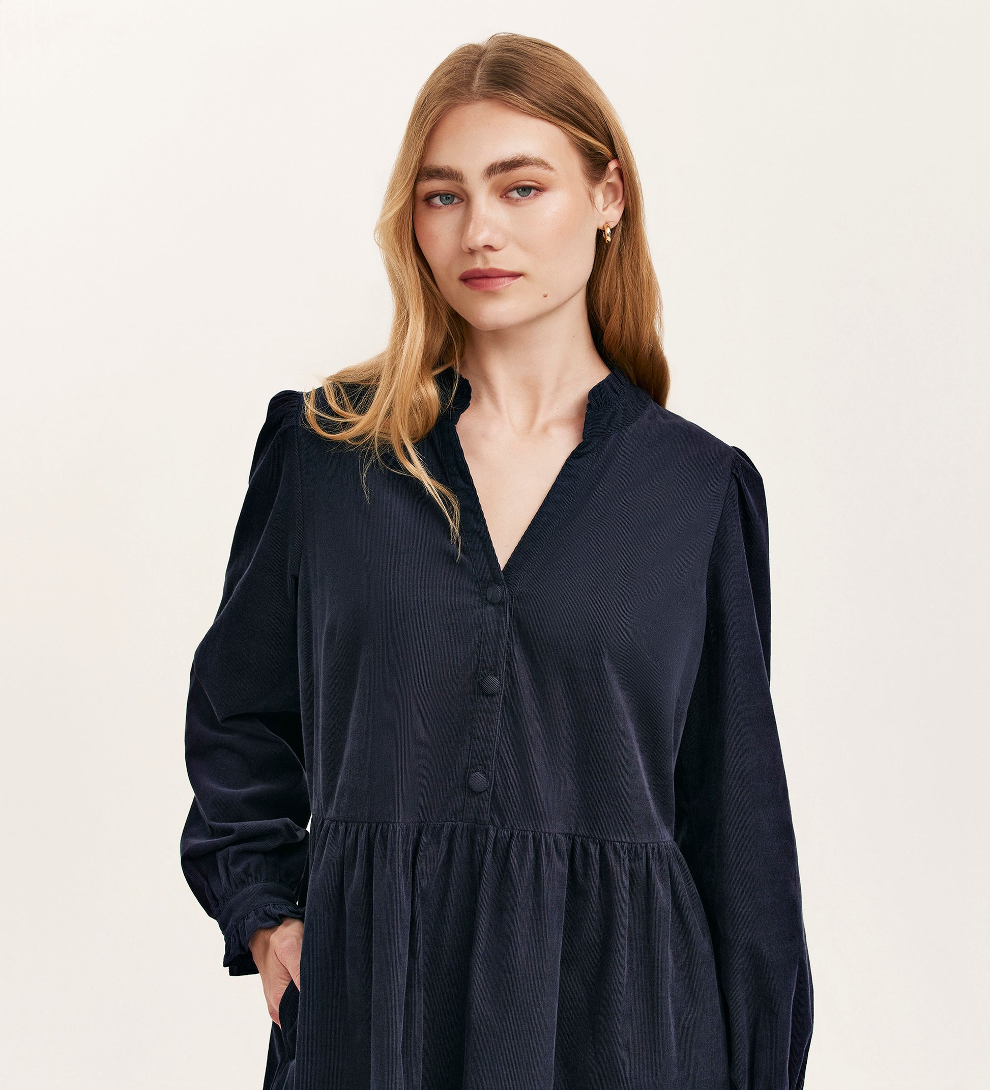 A woman with long blonde hair wears the Verona Navy Corduroy Short Dress, featuring a v-neck and buttons, standing against a plain light background and looking directly at the camera with a neutral expression.