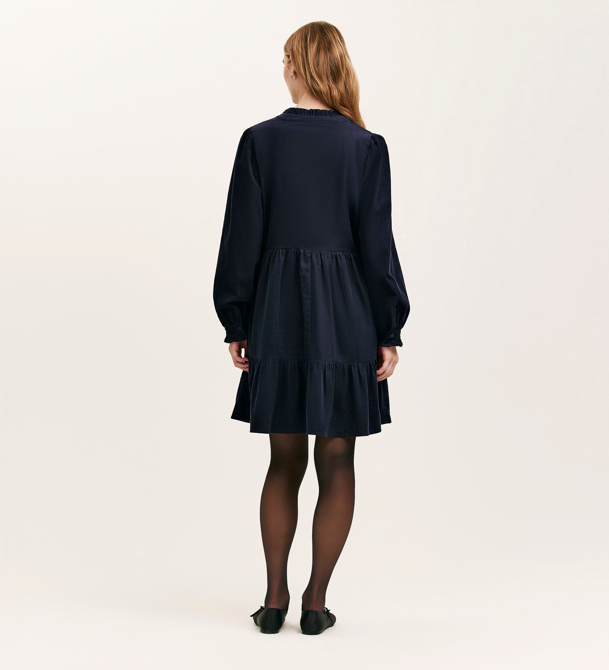 A woman with light brown hair wears the Verona Navy Corduroy Short Dress featuring long sleeves and a tiered skirt, paired with black tights and shoes. She stands facing away against a plain, light-colored background.