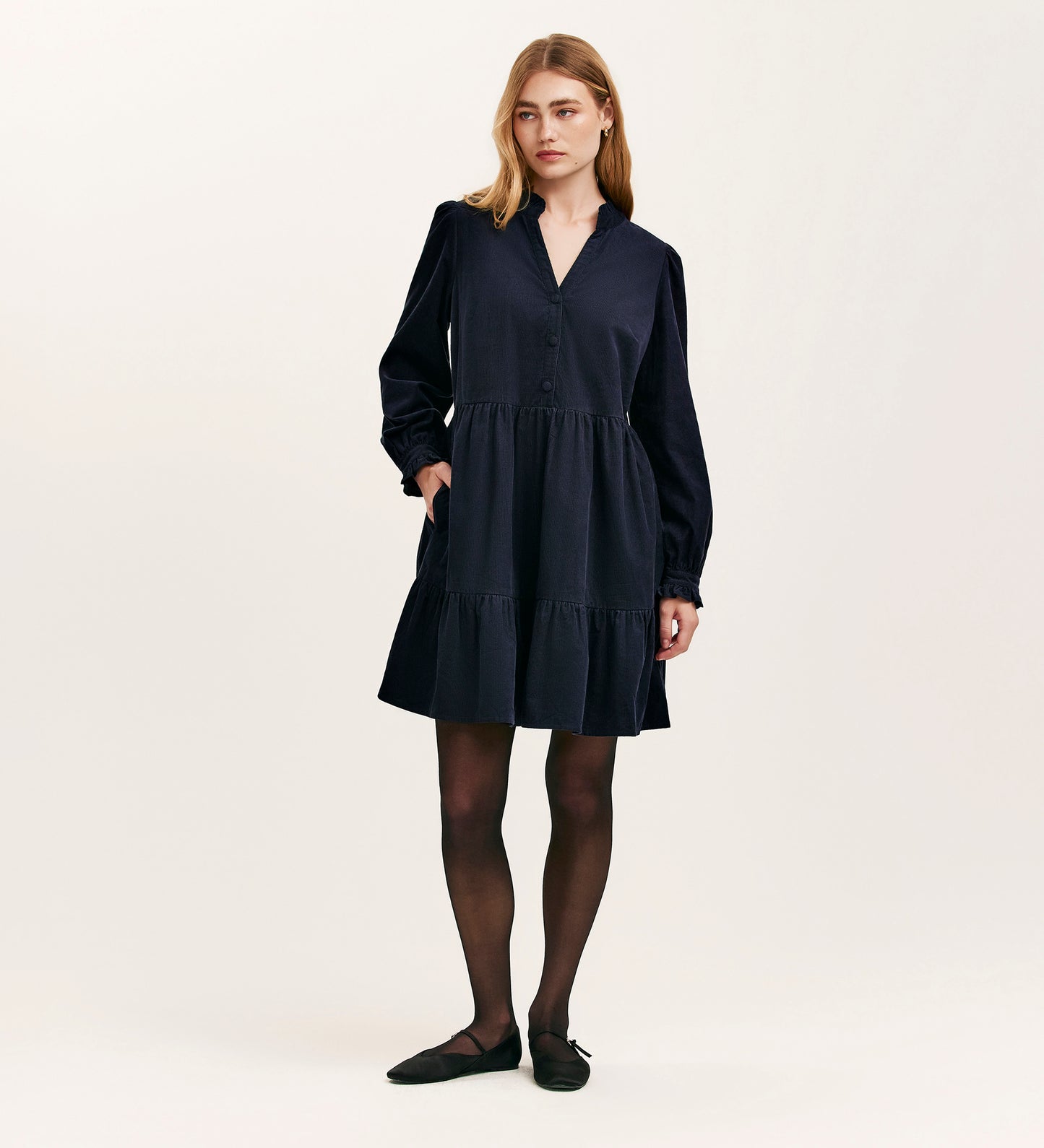 A woman with shoulder-length blonde hair and a neutral expression stands against a plain, light background wearing the Verona Navy Corduroy Short Dress, paired with black tights and flats.