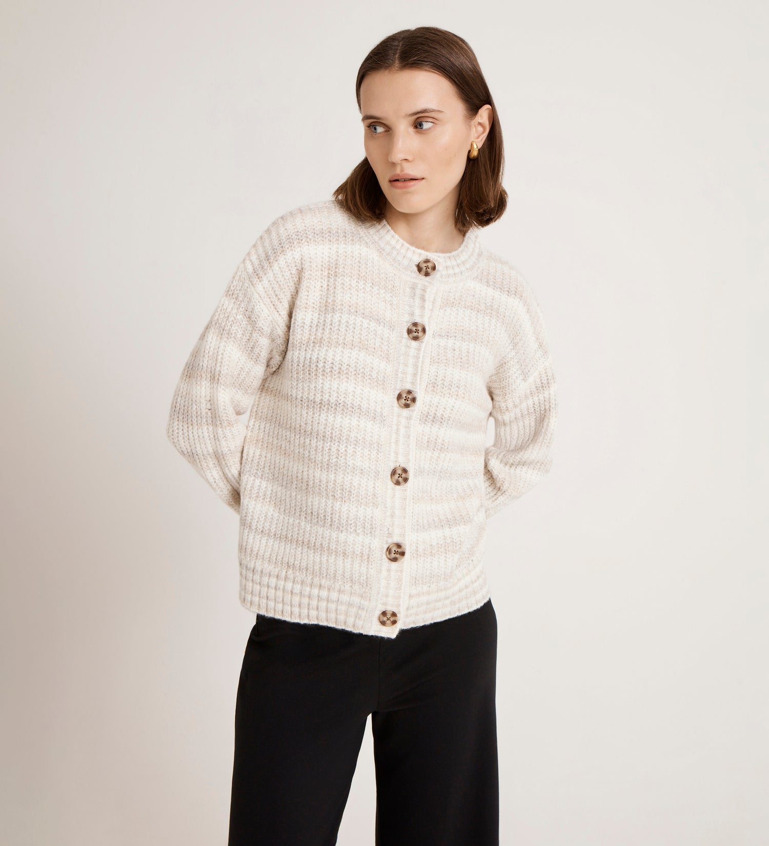 A woman with straight brown hair wears the Whitney Ivory Stripe Knitted Cardigan with mock horn buttons and black pants, standing against a plain white background, hands behind her back and gazing slightly to the side.