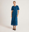 A person stands against a plain light background, wearing the Yasmine Blue Mesh Midi Tea Dress featuring a relaxed fit, lace details, fitted waist, and ruffled hem. Their light hair is pulled back and they’re wearing black flats.