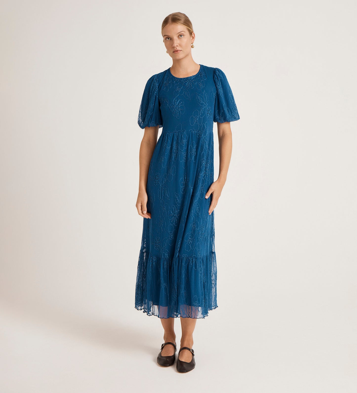 A person stands against a plain light background, wearing the Yasmine Blue Mesh Midi Tea Dress featuring a relaxed fit, lace details, fitted waist, and ruffled hem. Their light hair is pulled back and they’re wearing black flats.
