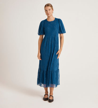 A person stands against a plain light background, wearing the Yasmine Blue Mesh Midi Tea Dress featuring a relaxed fit, lace details, fitted waist, and ruffled hem. Their light hair is pulled back and they’re wearing black flats.