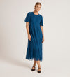 A woman in the Yasmine Blue Mesh Midi Tea Dress, featuring short sleeves, a tiered skirt, and ruffled hem, stands against a light background with her hair pulled back and black Mary Jane shoes.