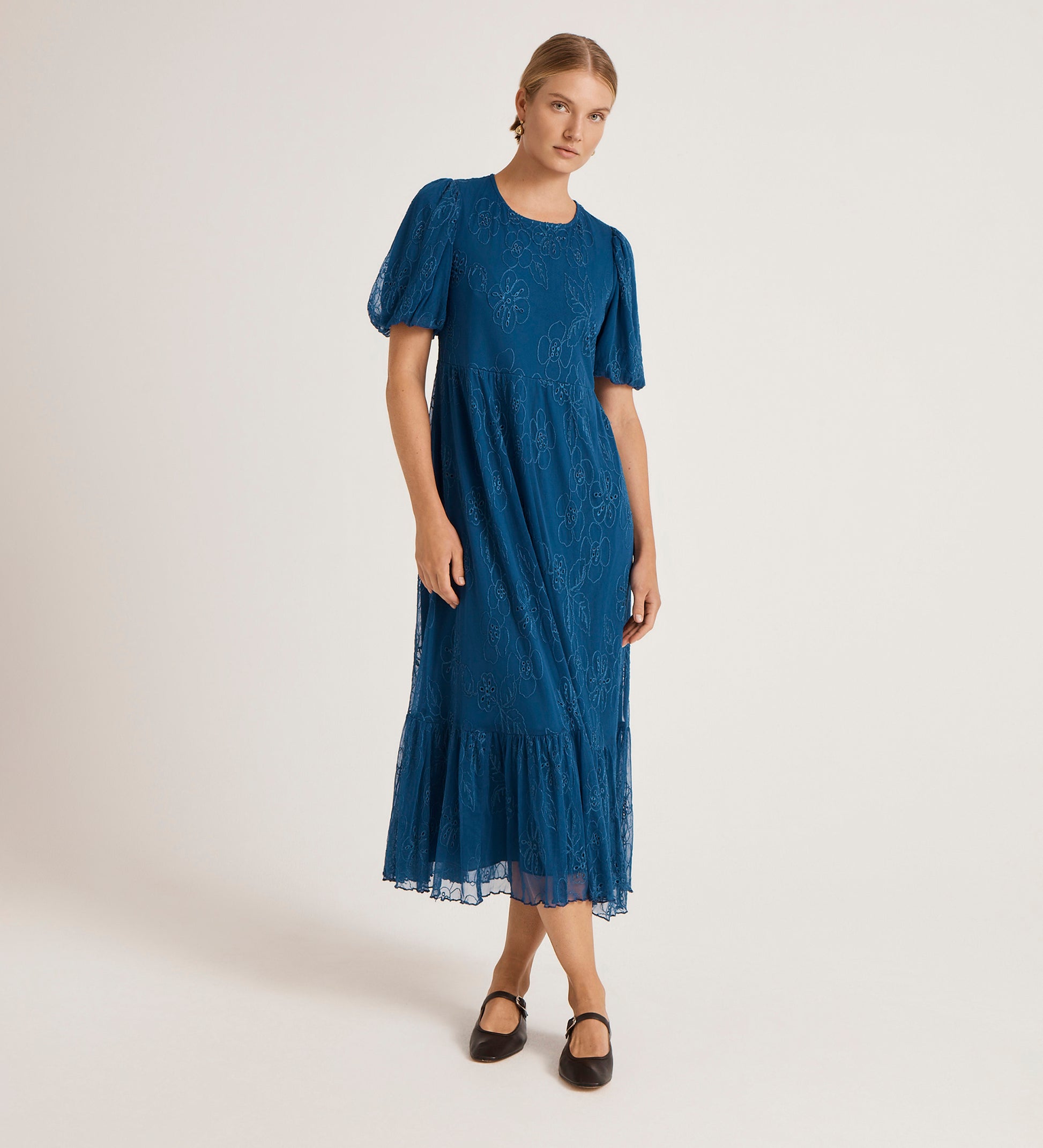 A woman in the Yasmine Blue Mesh Midi Tea Dress, featuring short sleeves, a tiered skirt, and ruffled hem, stands against a light background with her hair pulled back and black Mary Jane shoes.