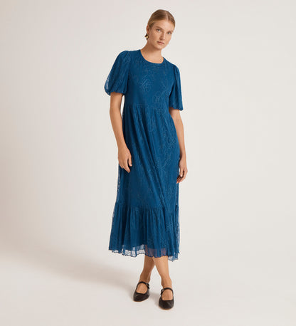 A woman in the Yasmine Blue Mesh Midi Tea Dress, featuring short sleeves, a tiered skirt, and ruffled hem, stands against a light background with her hair pulled back and black Mary Jane shoes.