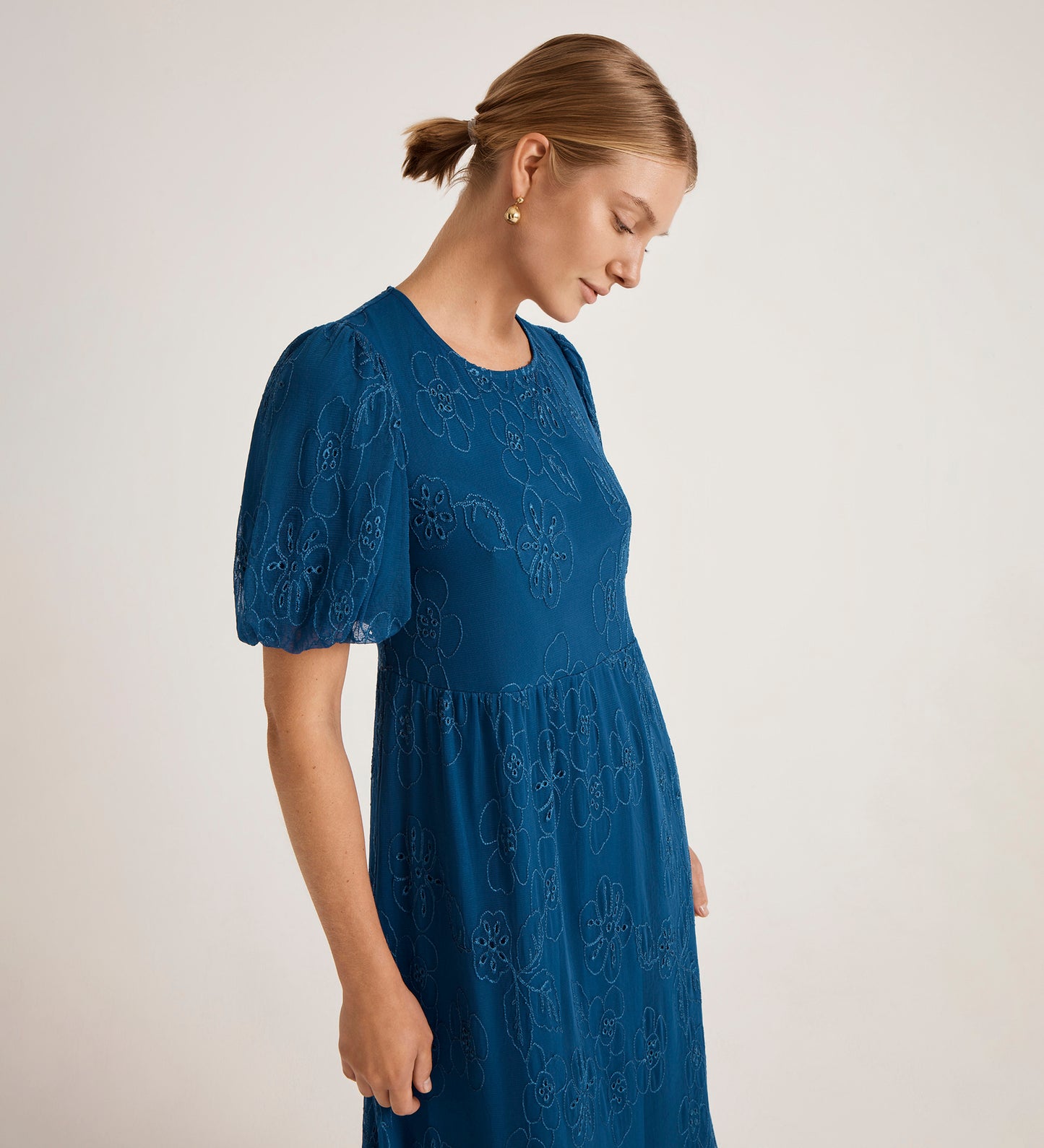 A woman with blonde hair in a low bun wears the Yasmine Blue Mesh Midi Tea Dress, featuring a relaxed fit, teal blue color, short sleeves, and a floral pattern. She looks down while standing against a plain light background.