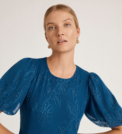A woman with blonde hair, wearing gold earrings and the Yasmine Blue Mesh Midi Tea Dress with floral embroidery, puffed sleeves, and a tiered skirt, stands against a plain light background.