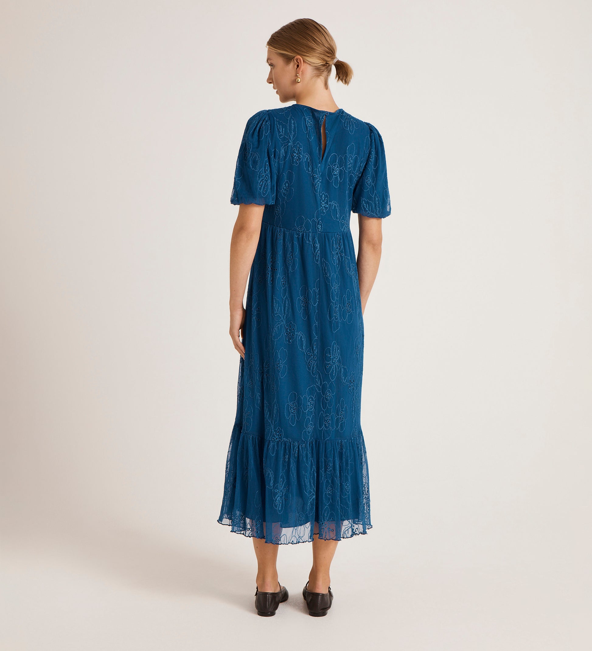 A woman with her hair tied back faces away, wearing the Yasmine Blue Mesh Midi Tea Dress—short-sleeved with floral embroidery, lace details, and a ruffled hem—paired with black shoes against a plain background.