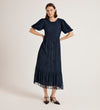 A woman stands with hands on hips wearing the Yasmine Navy Mesh Midi Tea Dress, which features puff sleeves, a subtle floral pattern, tiered skirt, and ruffle hem. She pairs it with black pointed-toe shoes against a plain light background.