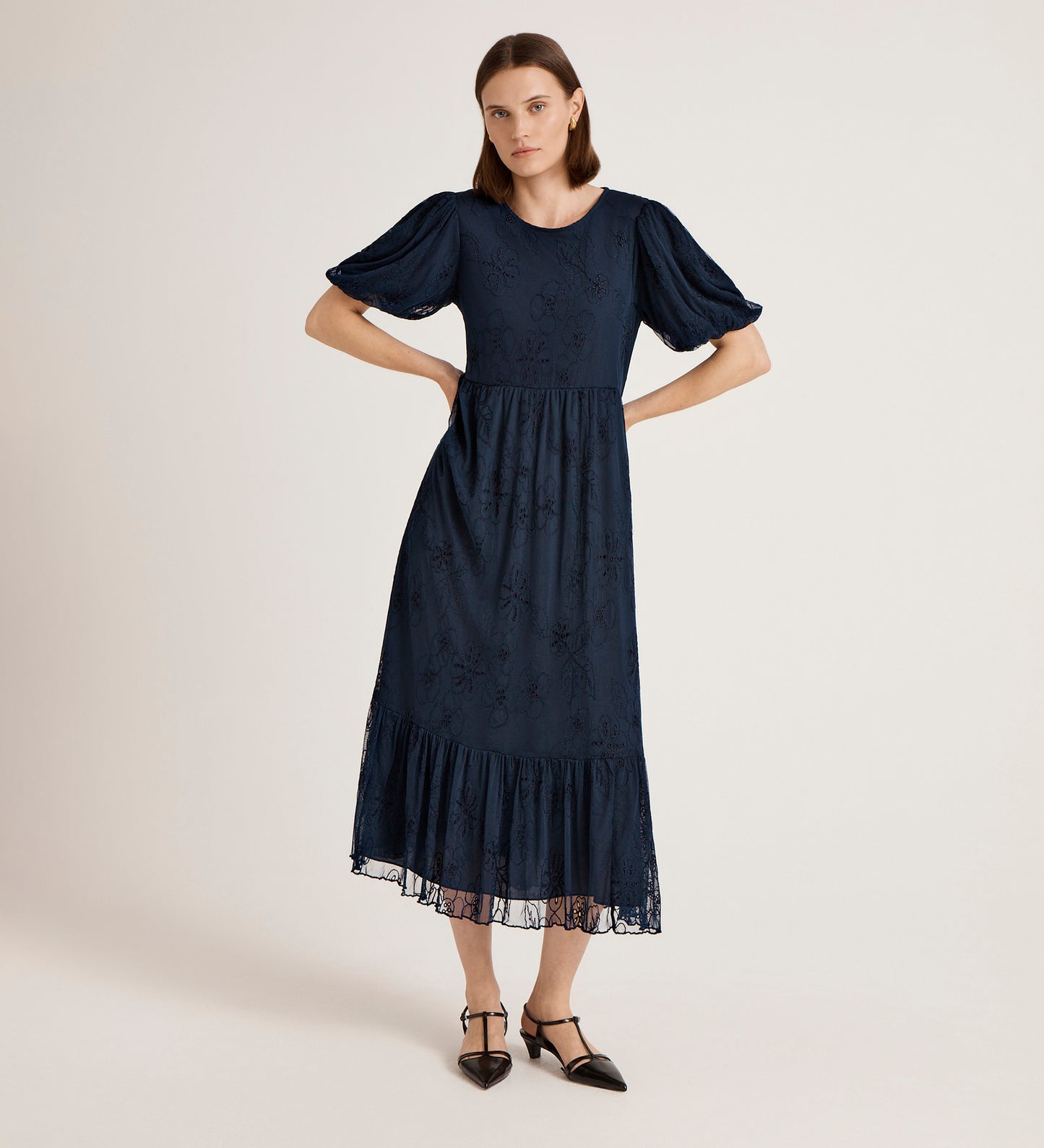 A woman stands with hands on hips wearing the Yasmine Navy Mesh Midi Tea Dress, which features puff sleeves, a subtle floral pattern, tiered skirt, and ruffle hem. She pairs it with black pointed-toe shoes against a plain light background.