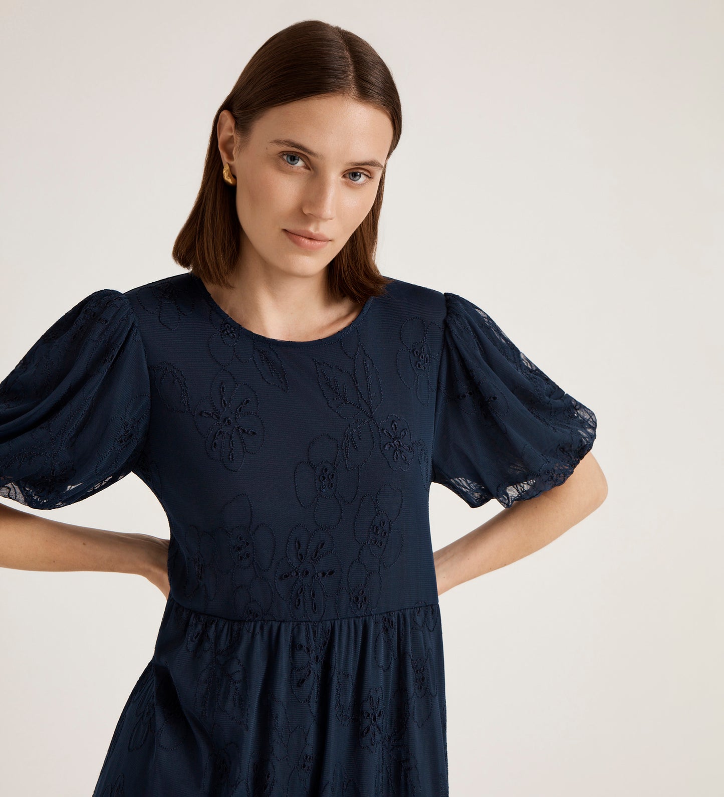 A woman with straight brown hair and gold hoop earrings wears the Yasmine Navy Mesh Midi Tea Dress, featuring floral embroidery and puff sleeves, standing against a plain light background with her hands on her hips.