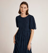 A woman with straight brown hair wears the Yasmine Navy Mesh Midi Tea Dress, featuring short sleeves and floral embroidery, standing against a plain light background.