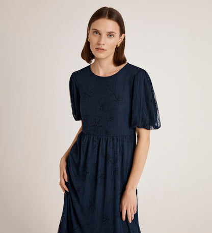A woman with straight brown hair wears the Yasmine Navy Mesh Midi Tea Dress, featuring short sleeves and floral embroidery, standing against a plain light background.