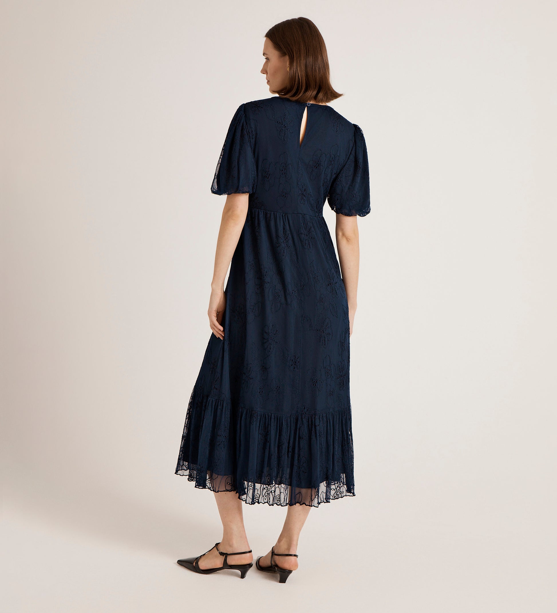 A woman with short brown hair stands facing away, modeling the Yasmine Navy Mesh Midi Tea Dress with puff sleeves, tiered skirt, and lace detail, paired with black low-heeled shoes against a plain light background.
