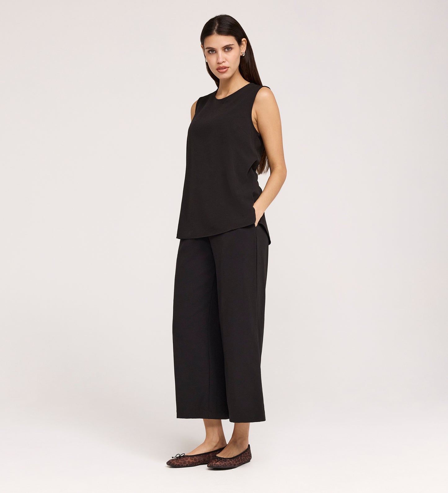 A woman with long dark hair and a neutral expression wears the Vallie Black Round Neck Vest and wide-leg black textured pants, hands in pockets, standing against a plain background with brown flat shoes.