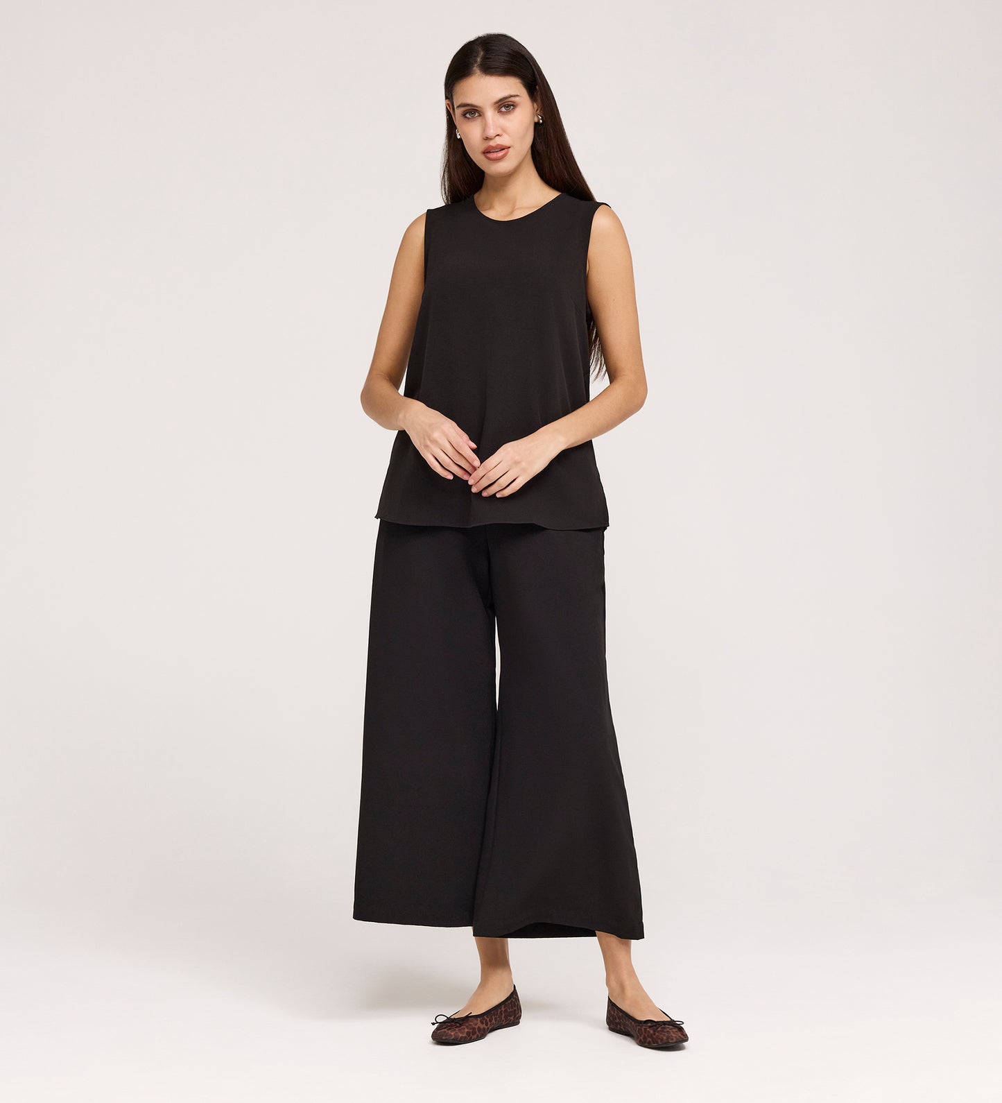 A woman with long dark hair and a neutral expression stands against a plain background, wearing the Vallie Black Round Neck Vest and loose, wide-leg black pants with dark flat shoes. Her hands rest in front of her.
