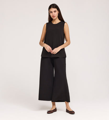 A woman with long dark hair and a neutral expression stands against a plain background, wearing the Vallie Black Round Neck Vest and loose, wide-leg black pants with dark flat shoes. Her hands rest in front of her.