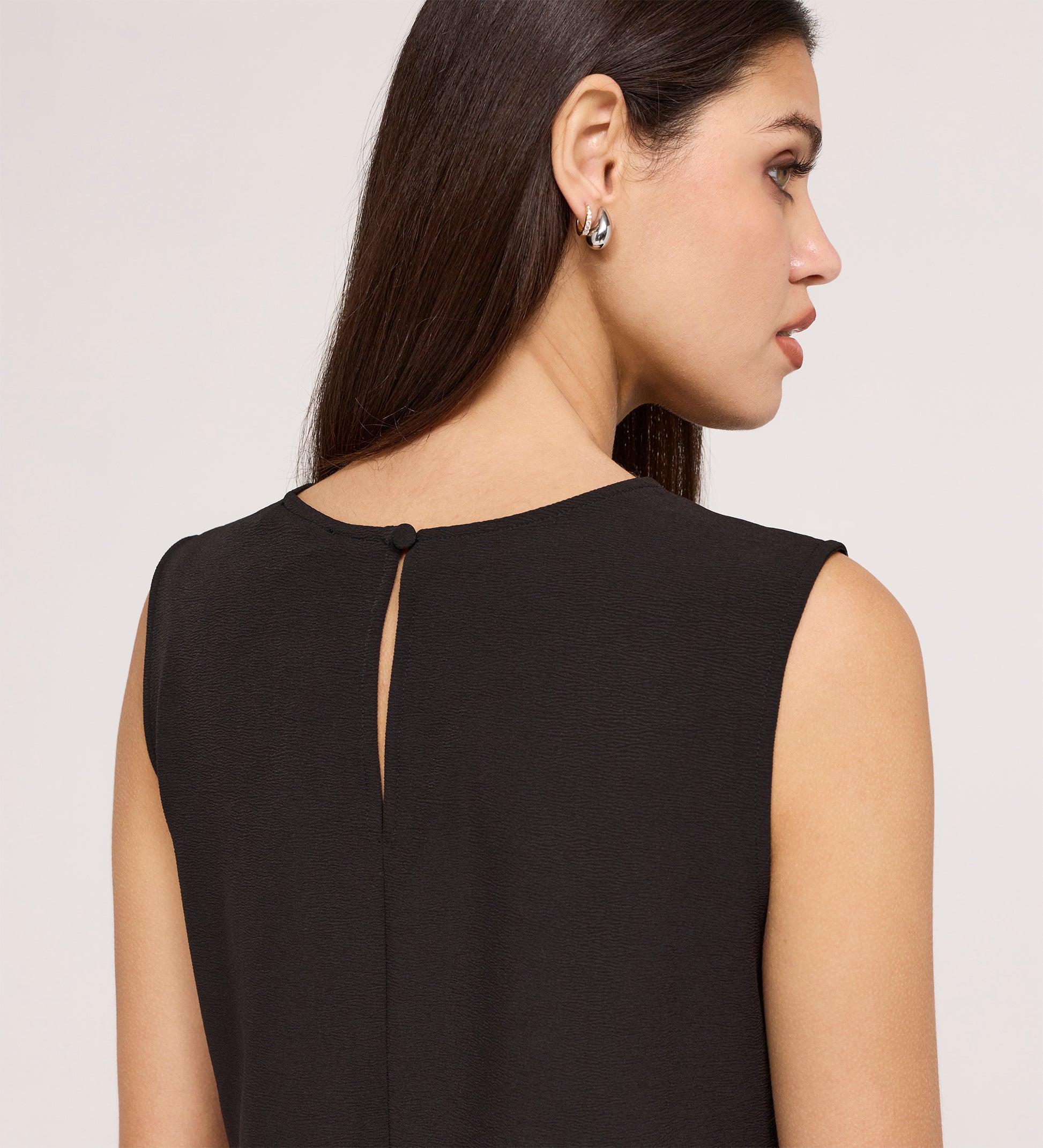 A woman with long brown hair is seen from behind wearing the Vallie Black Round Neck Vest, featuring a sleeveless design, buttoned keyhole back, relaxed fit, and textured poly crinkle fabric. She pairs it with silver earrings.