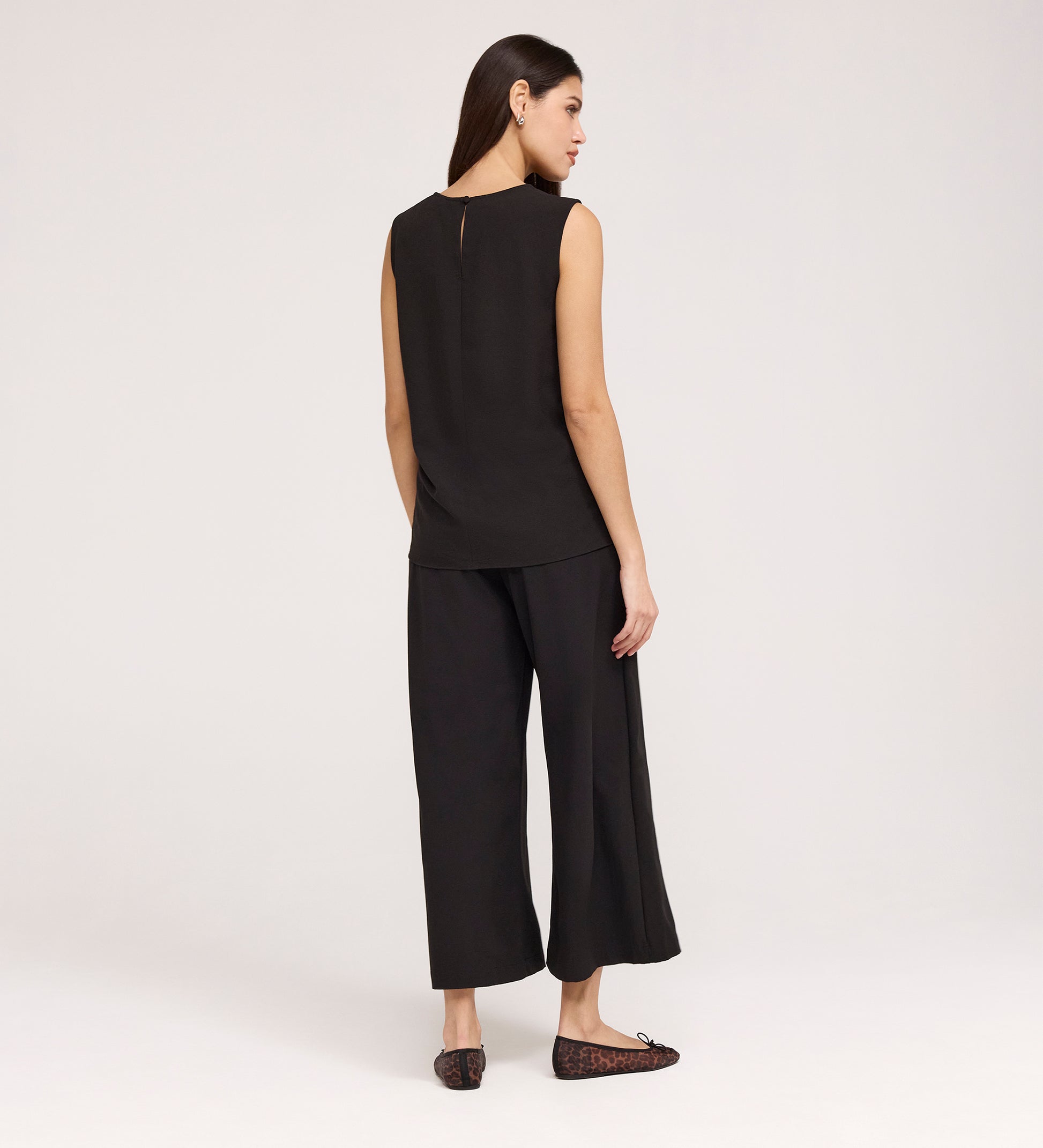 A woman faces away, wearing the Vallie Black Round Neck Vest and wide-leg black pants in textured poly crinkle with black flats, set against a plain light background.