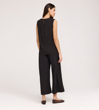 A woman faces away, wearing the Vallie Black Round Neck Vest and wide-leg black pants in textured poly crinkle with black flats, set against a plain light background.