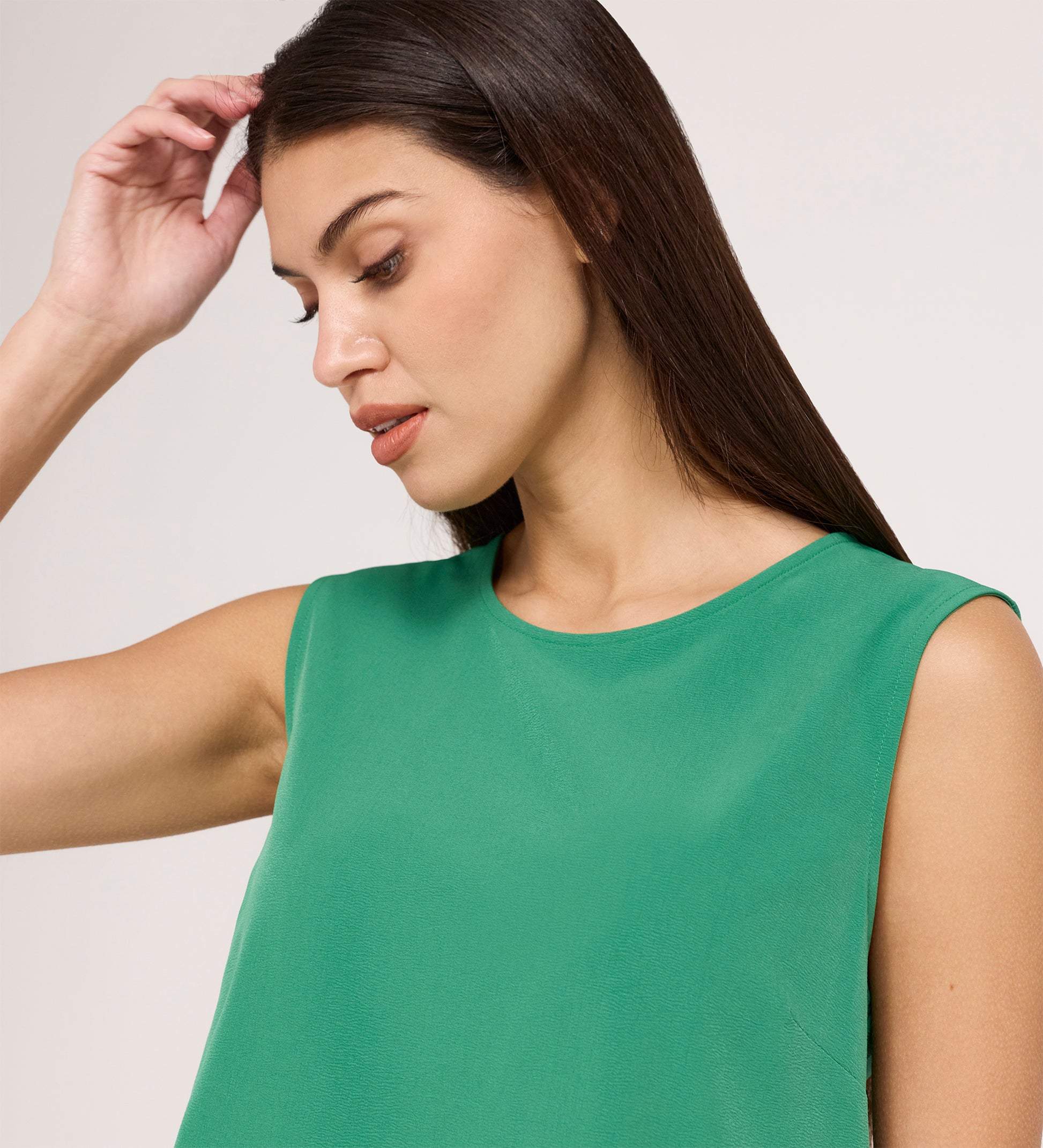 A woman with long dark hair wears the Vallie Green Round Neck Vest—a relaxed fit, sleeveless top—and looks down thoughtfully with one hand raised near her head against a plain background.