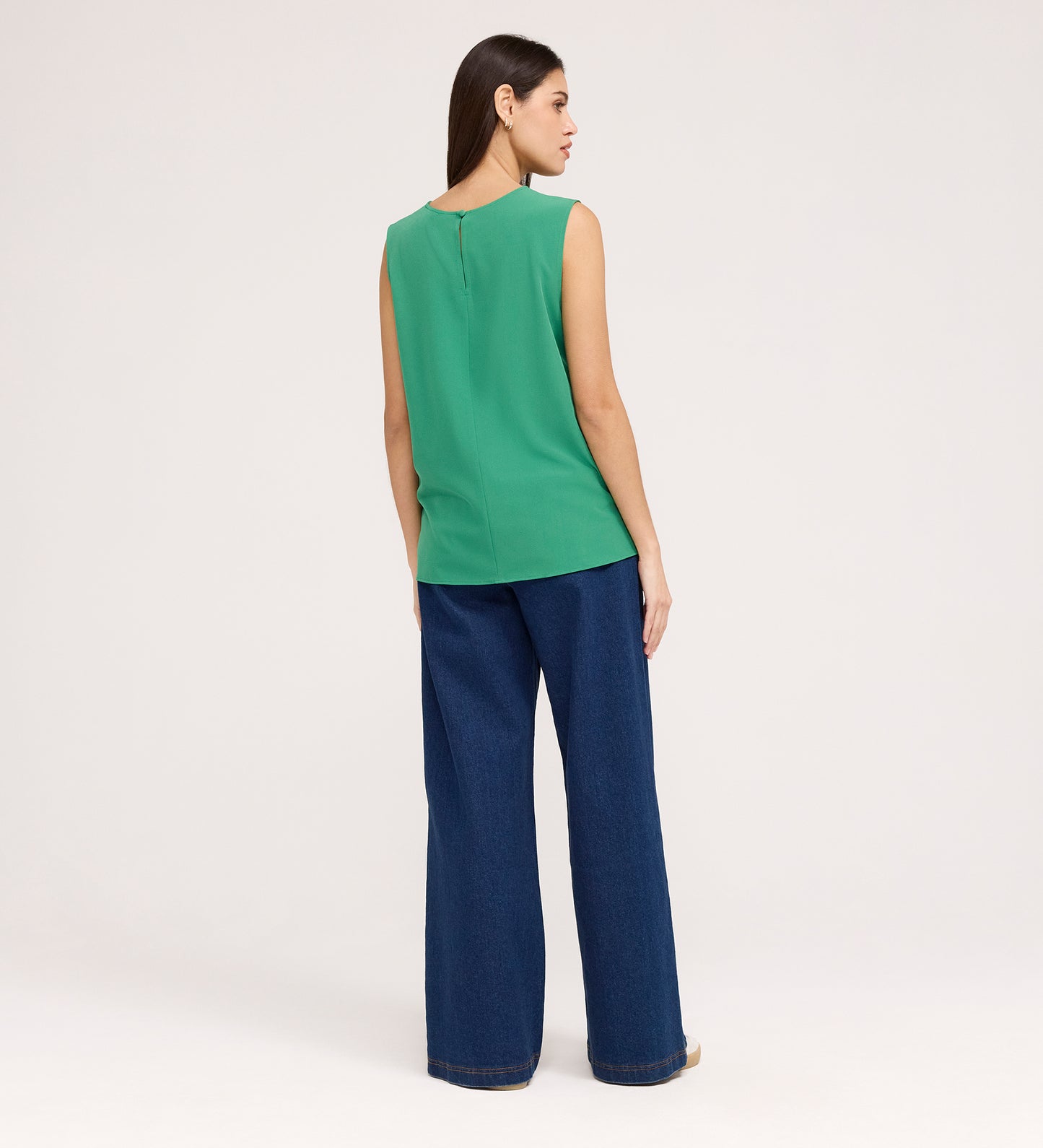 A woman with straight brown hair stands facing away, wearing the Vallie Green Round Neck Vest and wide-leg blue jeans against a plain light background.
