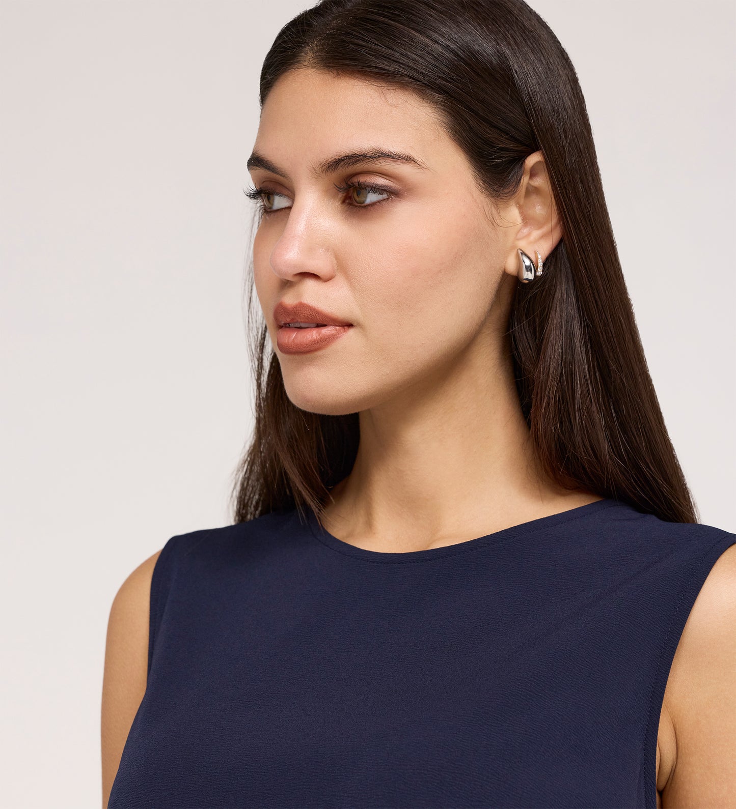 A woman with long, straight brown hair wears the Vallie Navy Round Neck Vest in textured poly crinkle and silver statement earrings, gazing to the side against a neutral background.