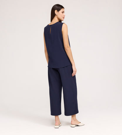 A woman stands facing away wearing the Vallie Navy Round Neck Vest and matching wide-leg cropped pants in textured navy poly crinkle, paired with light flat shoes, against a plain light background.