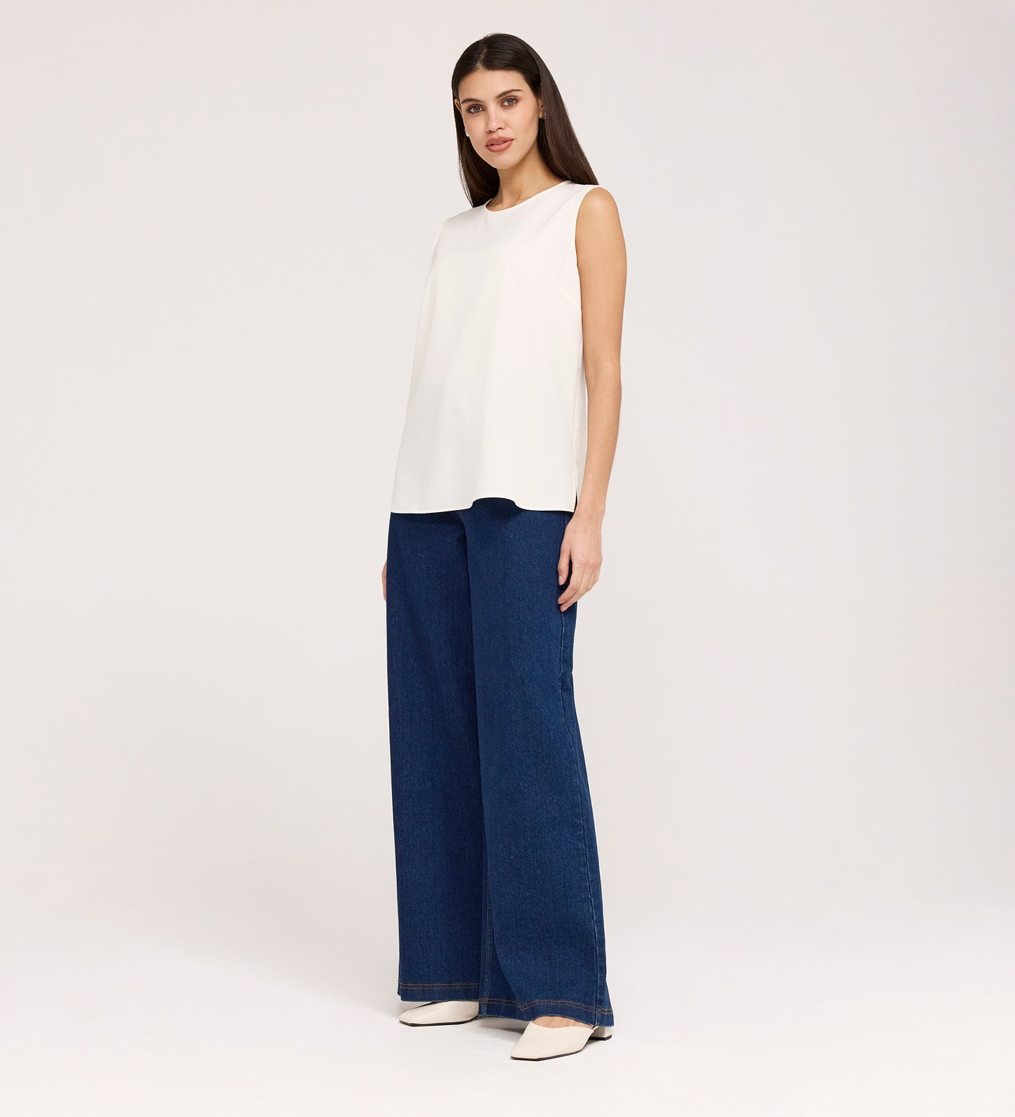 A woman stands against a plain background wearing the Vallie Ivory Round Neck Vest, relaxed-fit wide-leg dark blue poly crinkle jeans, and white pointed-toe shoes; her brown hair is down and her hands are by her sides.