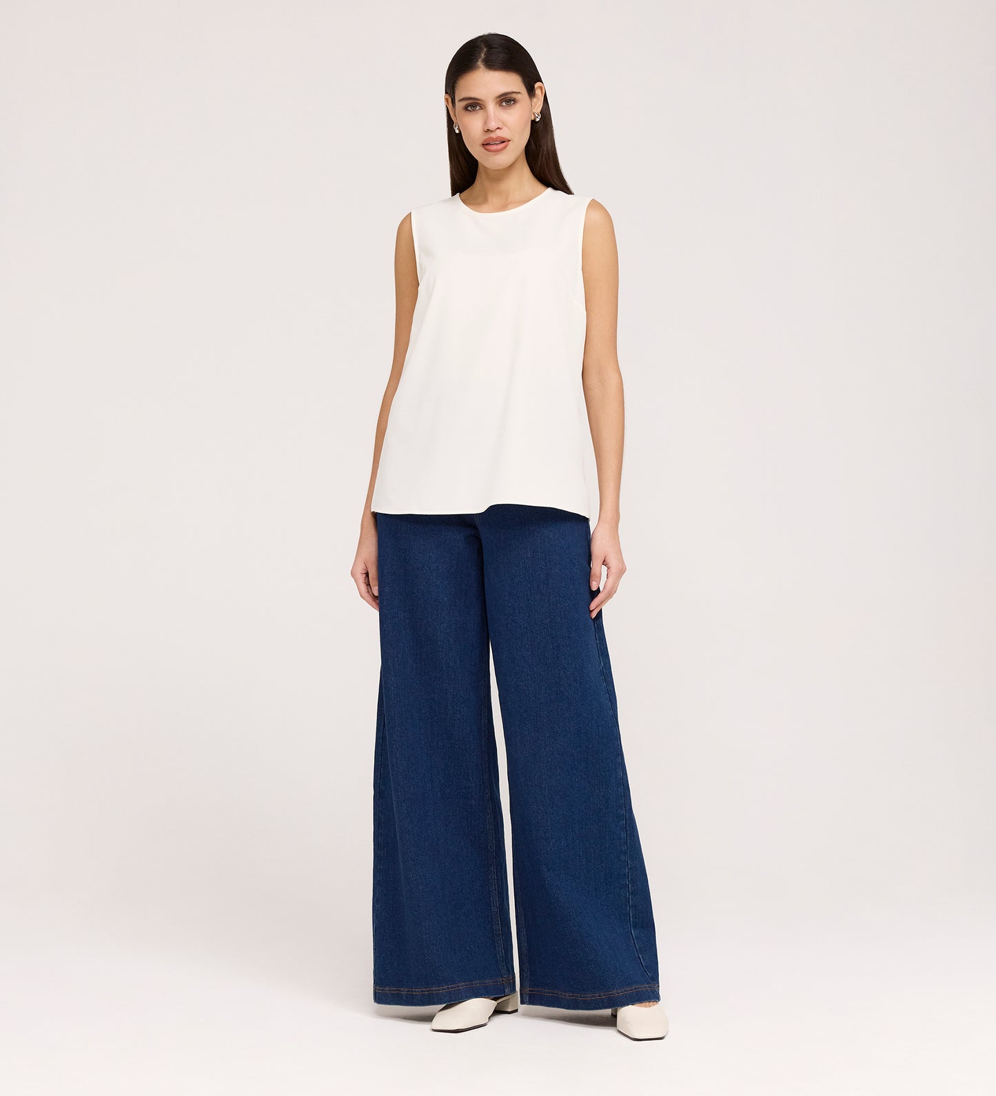 A woman stands facing forward, wearing the Vallie Ivory Round Neck Vest and high-waisted, wide-leg blue jeans with a relaxed fit against a plain white background.