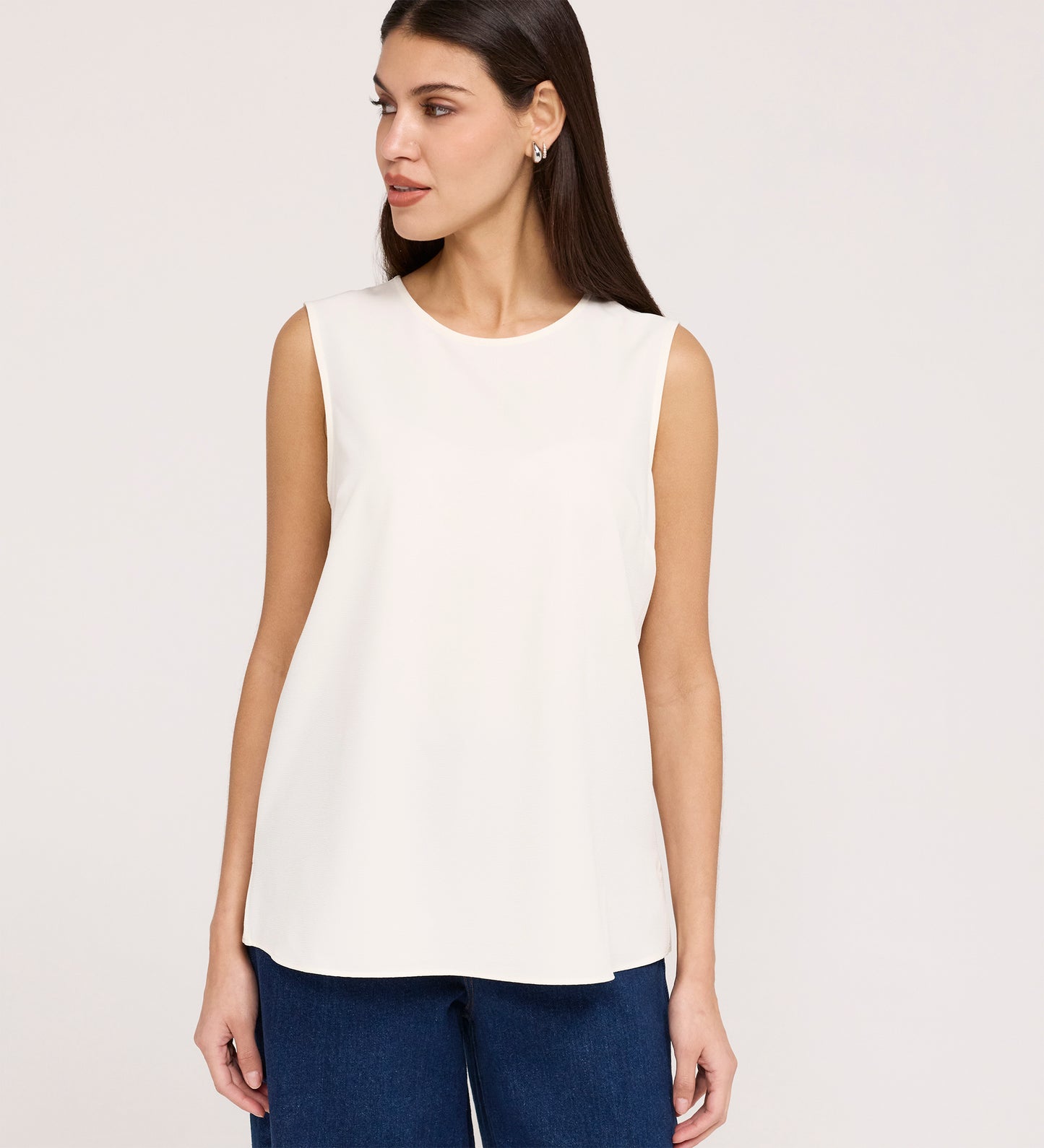 A woman with straight brown hair, wearing the Vallie Ivory Round Neck Vest and blue jeans, stands against a plain light background, looking to her left with a neutral expression.