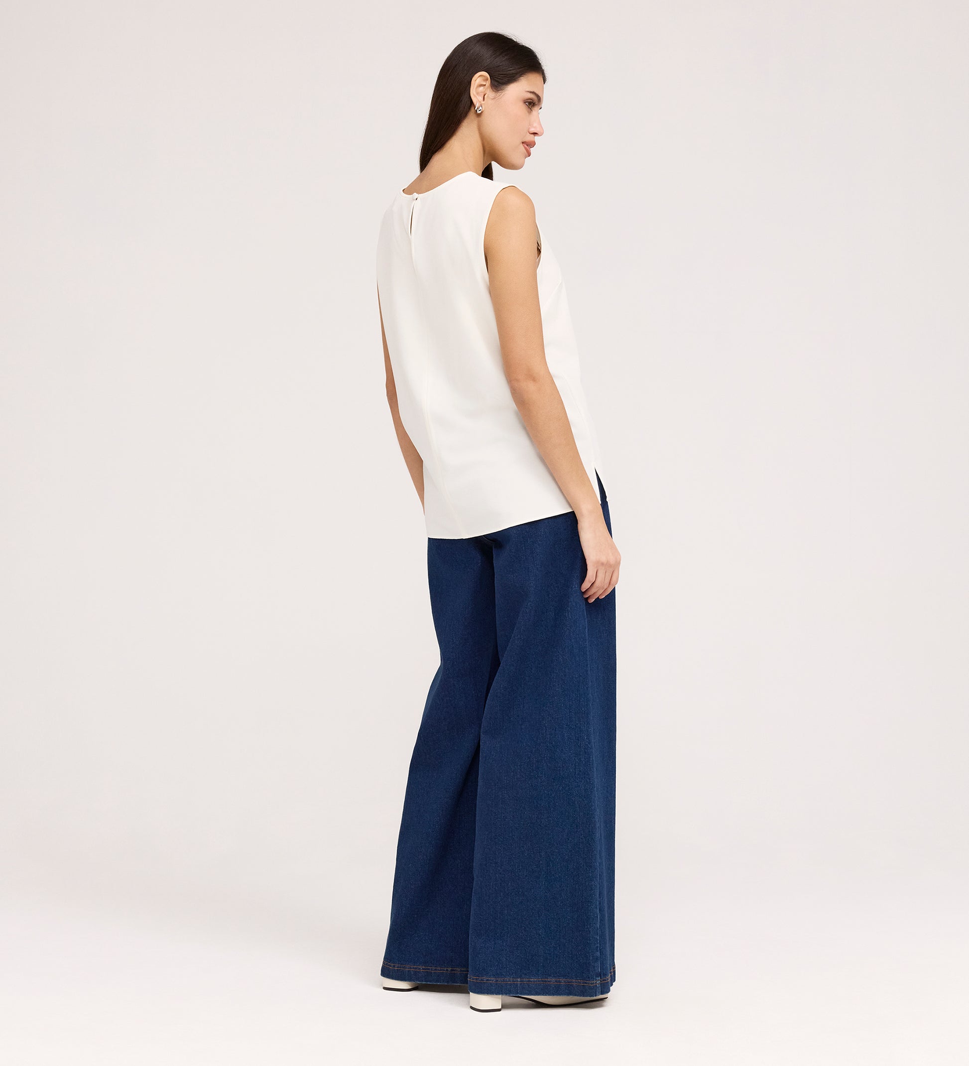 A woman stands facing slightly away, wearing the Vallie Ivory Round Neck Vest with wide-leg blue jeans and white shoes against a plain light background.